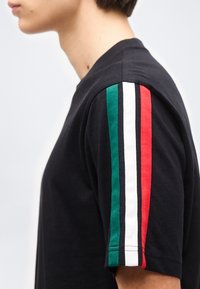 Külgvaade inimesest, kes kannab musta lühikeste varrukatega särk, mille varrukal on rohelised, valged ja punased vertikaalsed triibud.