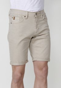 Beige rullade denimshorts bärs av en person med synliga ben och en vit skjorta instoppad mot en neutral bakgrund.