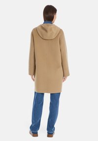 Camel-farbener Mantel mit Kapuze, aus weichem Stoff gefertigt, mit geradem Schnitt und langen Ärmeln. Wird über blauen Jeans getragen.