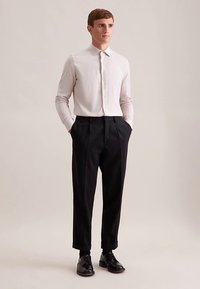 Camicia grigia chiaro a coste con colletto e chiusura con bottoni, abbinata a pantaloni neri plissettati e scarpe di pelle nere.