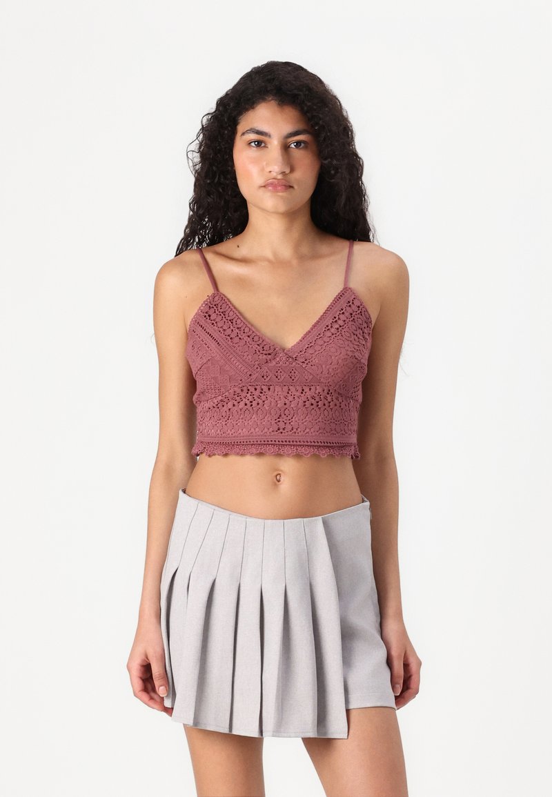 Kvačkana crop majica u prašnjavo ružičastoj boji s V-izrezom i valovitim rubom, uparena s sivom plisiranom A-linijom mini suknjom. Glatke teksture kroz cijeli outfit.