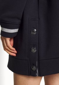 Mörk marinblå sweatshirt med textilstruktur, ribbad mudd med silverrand och dekorativa svarta knappar på sidan.