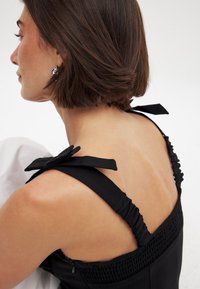 Vestito nero con spalline arricciate e dettagli a fiocco. Presenta particolari smock e una texture di tessuto liscia. Cerniera laterale visibile.