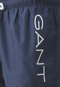 Jūriniai mėlynos spalvos maudymosi šortai su elastinga juosta ir balta virvele. Šone vertikaliai spausdintas didelis baltas logotipas.