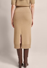 Strickmidirock in Beige mit geripptem Saum und einem Schlitz hinten, kombiniert mit knielangen braunen Stiefeln. Weiche Textur, figurbetonte Passform.