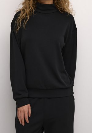 Frau trägt einen schwarzen Rollkragenpullover mit langen Ärmeln und passende schwarze Hosen, steht vor einem einfachen weißen Hintergrund.