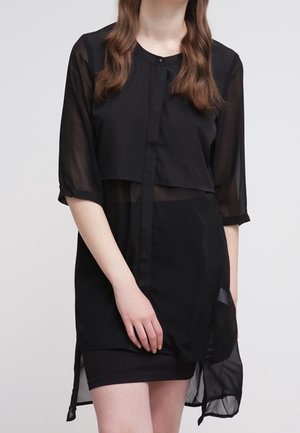 Femme portant un long chemisier noir transparent à manches trois-quarts par-dessus une robe noire sans manches, debout les mains jointes devant elle.