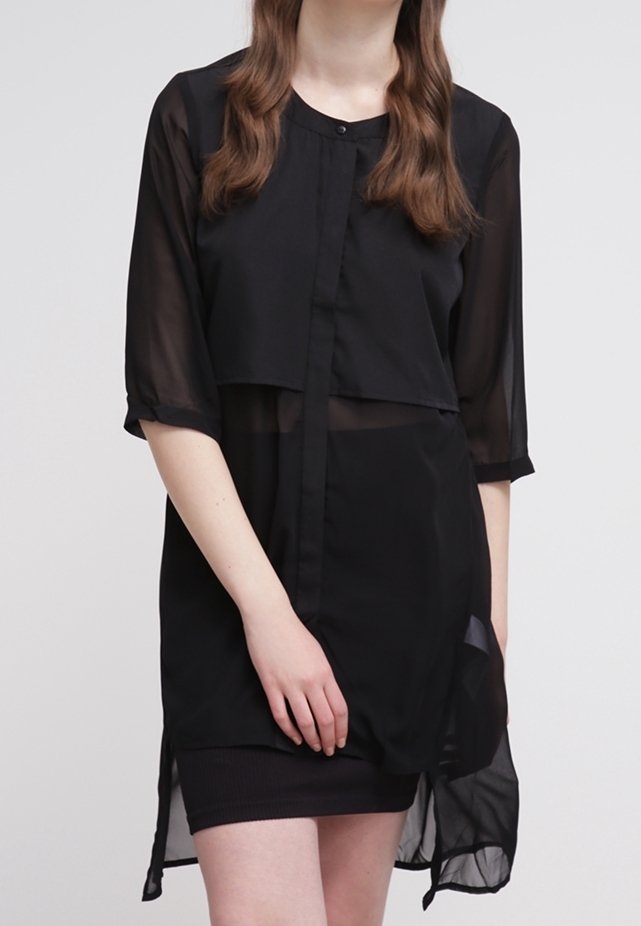 Femme portant un long chemisier noir transparent à manches trois-quarts par-dessus une robe noire sans manches, debout les mains jointes devant elle.
