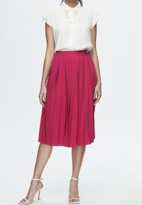 Gonna plissettata fucsia con vita alta, abbinata a una blusa bianca a maniche corte. La modella indossa tacchi bianchi con cinturini. Tessuto dalla texture liscia.