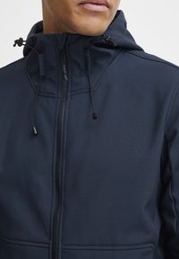 Marineblå hettegenser laget av myk, teksturert stoff, med hel glidelås, justerbare snorer og minimale sømdetaljer.