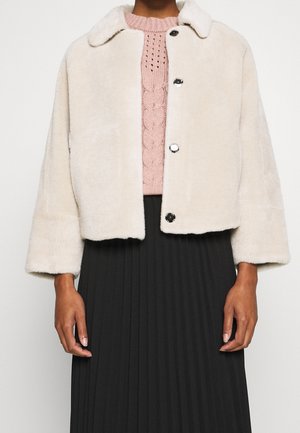 Mujer vistiendo una chaqueta peluda color crema sobre un suéter de punto rosa y una falda negra plisada sobre un fondo claro liso.