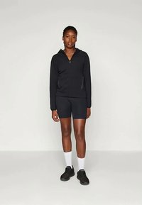 Fekete zippzáras kapucnis pulcsi oldalsó zsebekkel, fekete rövidnadrággal párosítva. A modellen fehér zoknik és fekete sportcipők vannak. Egyszerű és testhezálló dizájn.