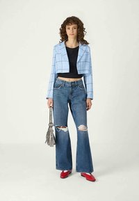 Jovem mulher com cabelo encaracolado, vestindo um blazer azul xadrez cortado, top preto, calças largas rasgadas e sapatos vermelhos, enquanto segura uma mala cinza.