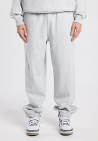 Pantalon de survêtement gris clair avec une taille élastique, des chevilles resserrées et une coupe décontractée, associé à des baskets blanches avec des accents gris.