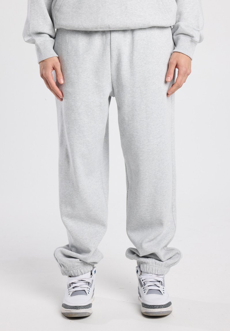 Pantalon de survêtement gris clair avec une taille élastique, des chevilles resserrées et une coupe décontractée, associé à des baskets blanches avec des accents gris.