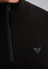 Pull zip noir avec col montant, fabriqué en tissu tricoté. Présente un léger côtelage et un petit logo brodé sur la poitrine.