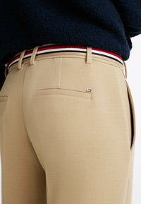 Khaki broek met een gladde structuur, voorzien van een contrasterende gestreepte riem in rood, wit en blauw, en een klein logodetail op de achterzak.