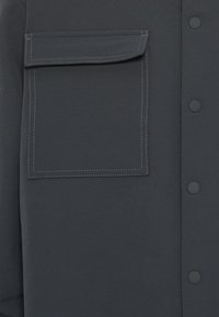 Dunkelgraues Hemd mit Knopfleiste und großer aufgesetzter Tasche. Verfügt über kontrastierende weiße Nähte entlang der Tasche und Knöpfe, glatte Stoffstruktur.