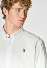 Camicia bianca con colletto, chiusura a bottoni, caratterizzata da un logo ricamato blu sul lato sinistro del petto. Tessuto di cotone liscio con una vestibilità classica.