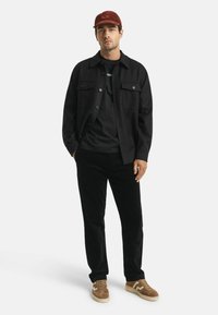 Man met een bordeauxrode pet, een zwarte button-up jack over een zwart shirt, zwarte broek en een paarse sneakers, staand met handen in zijn zakken.