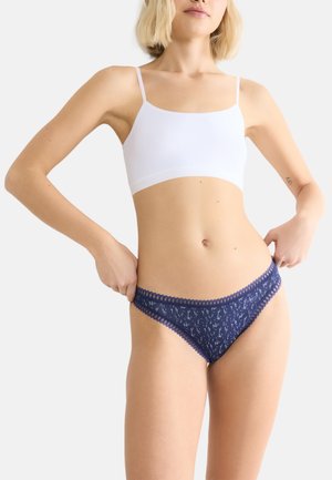 Witte bralette met dunne bandjes, gecombineerd met donkerblauwe panties met kantafwerking, voorzien van een subtiel wit patroon en golvende randen.