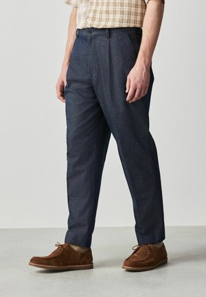 Mężczyzna ubrany w ciemnoniebieskie, dopasowane spodnie, beżową koszulę w kratę z krótkim rękawem oraz brązowe zamszowe buty wiązane, stojący na jasnoszarej podłodze.