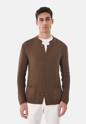 Giovane uomo che indossa un cardigan marrone a maglia con bottoni e due tasche frontali sopra una camicia bianca e pantaloni bianchi, in piedi davanti a uno sfondo chiaro.