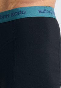 Crni bokserice od glatkog materijala. Pojas je tirkizne boje s ljubičastim natpisom "BJÖRN BORG". Šavne detalje duž strana.