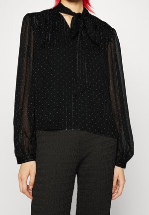 Femme portant un chemisier noir transparent à pois avec manches longues et un nœud au cou, associé à un pantalon foncé texturé.