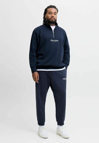 Mörkblå kvartzip-tröja med vit "Jack & Jones" logotyp på bröstet, matchad med joggers och vita sneakers.