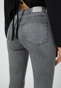 Jeans de denim gris con un ajuste slim, que presentan bolsillos traseros y una etiqueta de marca en la cinturilla. El material tiene un acabado suave y ligeramente texturizado.