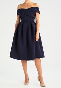 Robe marine décolletée épaules avec un corsage ajusté et une jupe plissée. Fabriquée en tissu lisse, avec un design croisé au niveau de l'encolure.