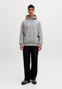 Nuori mies seisoo päällään harmaa huppari, jossa on logo, mustat housut ja harmaat lenkkarit valkoista taustaa vasten.