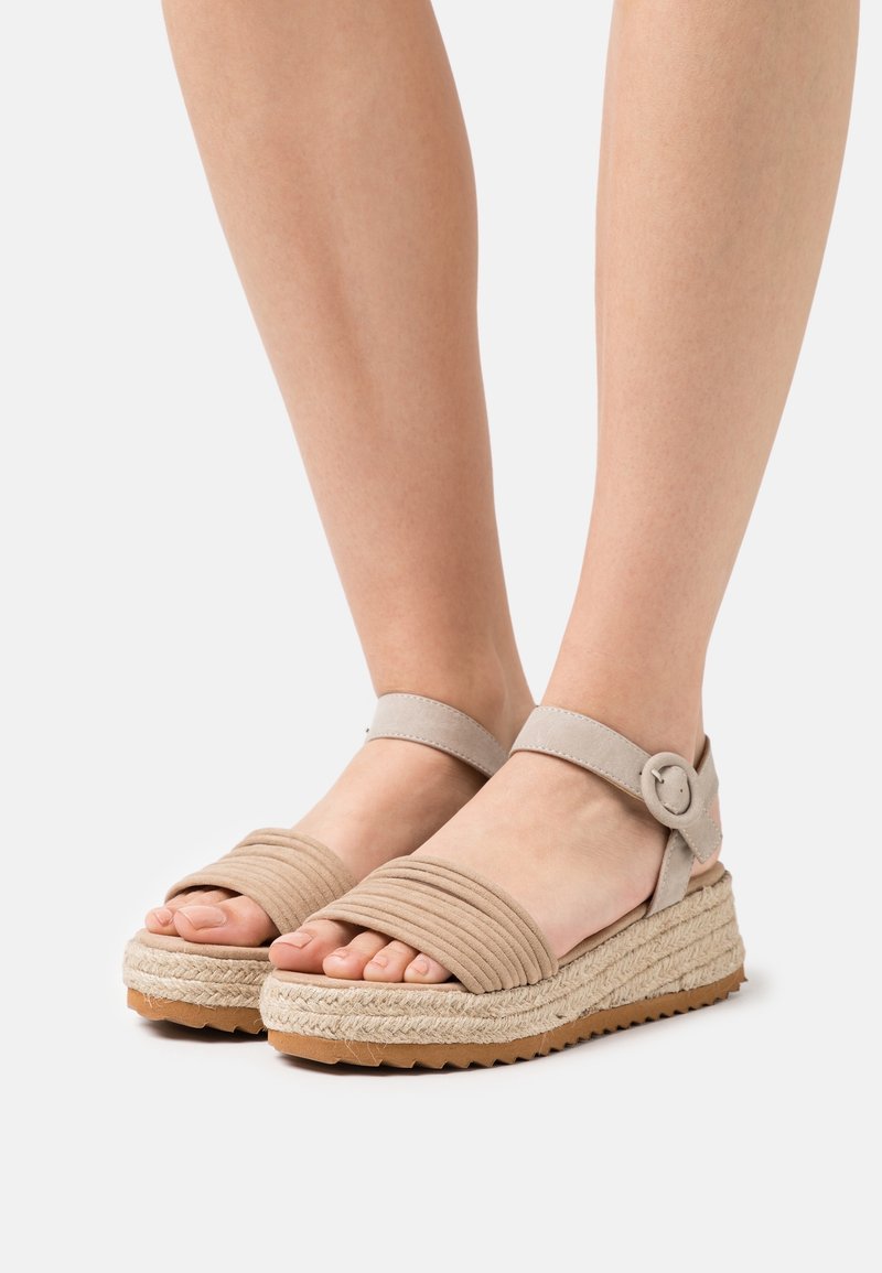 Sandales espadrilles beiges avec des brides épaisses et texturées. Présentent une boucle ronde sur une bride de cheville et une semelle en jute enveloppée.