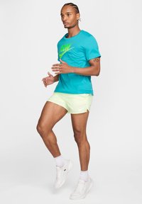 Camiseta Nike turquesa con gráfico de 'RUN', combinada con pantalones cortos amarillos claros. Zapatillas y calcetines blancos, postura atlética, textura de piel suave.