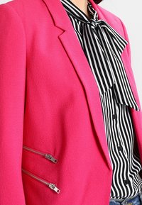 Fuchsia blazer i texturerad tyg med skräddarsydd design. Funktionen har silvriga dragkedjeaccentuerade fickor och en blus i svart och vitt ränder under.