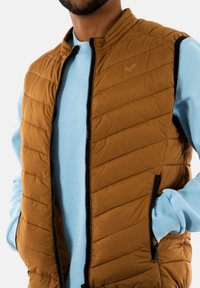 Gilet imbottito marrone con texture a quadri orizzontali, zip frontale completa e rifiniture nere. Indossato sopra una felpa azzurro chiaro.