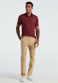 Man in een rood gestreept polo-shirt, beige broek en witte sneakers, staande met één hand in zijn zak tegen een eenvoudige achtergrond.