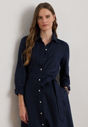 Femme aux longs cheveux ondulés portant une robe chemise bleu marine avec ceinture à la taille et manches retroussées, debout devant un mur clair uni.