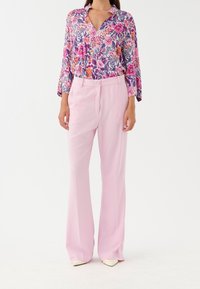 Blouse fleurie en rose vif et violet avec manches longues, associée à un pantalon à jambes larges rose clair et des chaussures blanches à bouts pointus.