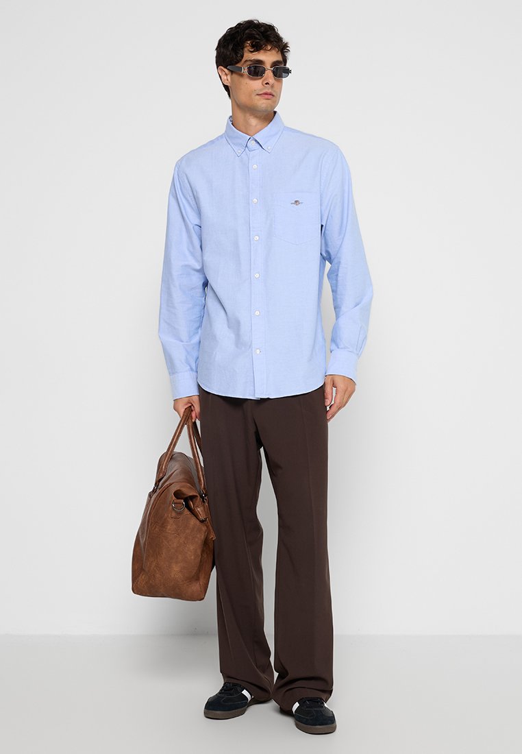 Jasnoniebieska koszula zapinana na guziki z długimi rękawami i kieszonką na piersi; zestawiona z brązowymi spodniami i brązową skórzaną torbą; czarne sneakersy.