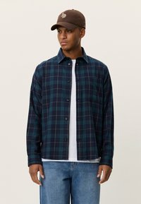 Φανελένιο πουκάμισο με καρό σχέδιο σε πράσινες και ναυτικές αποχρώσεις, κλείσιμο με κουμπιά, γιακά και τσέπη στο στήθος. Φορεμένο πάνω από ένα λευκό t-shirt.