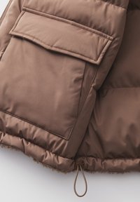 Braune Puffjacke mit einer großen Fronttasche, glattem Stoff und einem Kordelzug am Saum zur Anpassung. Texturierter Innenstoff sichtbar.