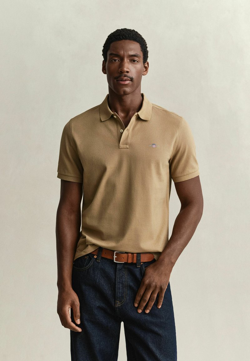 Homme portant un polo beige à manches courtes, un jean foncé et une ceinture en cuir marron, debout devant un fond clair uni.