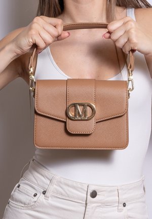 Femme en haut blanc tenant un sac à main en cuir fauve avec fermoir doré centré devant son torse sur fond neutre.