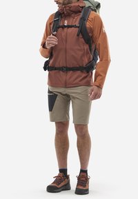 Veste d'extérieur en rouille avec un col montant ; associée à un short beige avec un panneau noir. Des chaussures de randonnée marron et un sac à dos vert complètent le look.