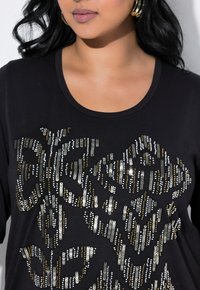 Acercamiento de una mujer que lleva una camiseta negra adornada con brillantes lentejuelas de varios colores, con un diseño decorativo audaz.