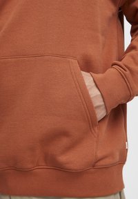 Sudadera color óxido con un bolsillo canguro, puños acanalados y dobladillo. Textura suave y diseño casual, con detalles de costura visibles.