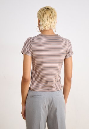 Personne aux cheveux blonds courts et boucles d'oreilles anneaux portant un t-shirt rayé marron et blanc ainsi qu'un pantalon gris, vue de dos.