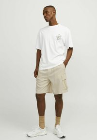 T-shirt blanche à col rond avec une impression graphique sur la poitrine gauche, accompagnée de shorts cargo beiges dotés de deux poches latérales et d'une coupe décontractée. Baskets blanches.